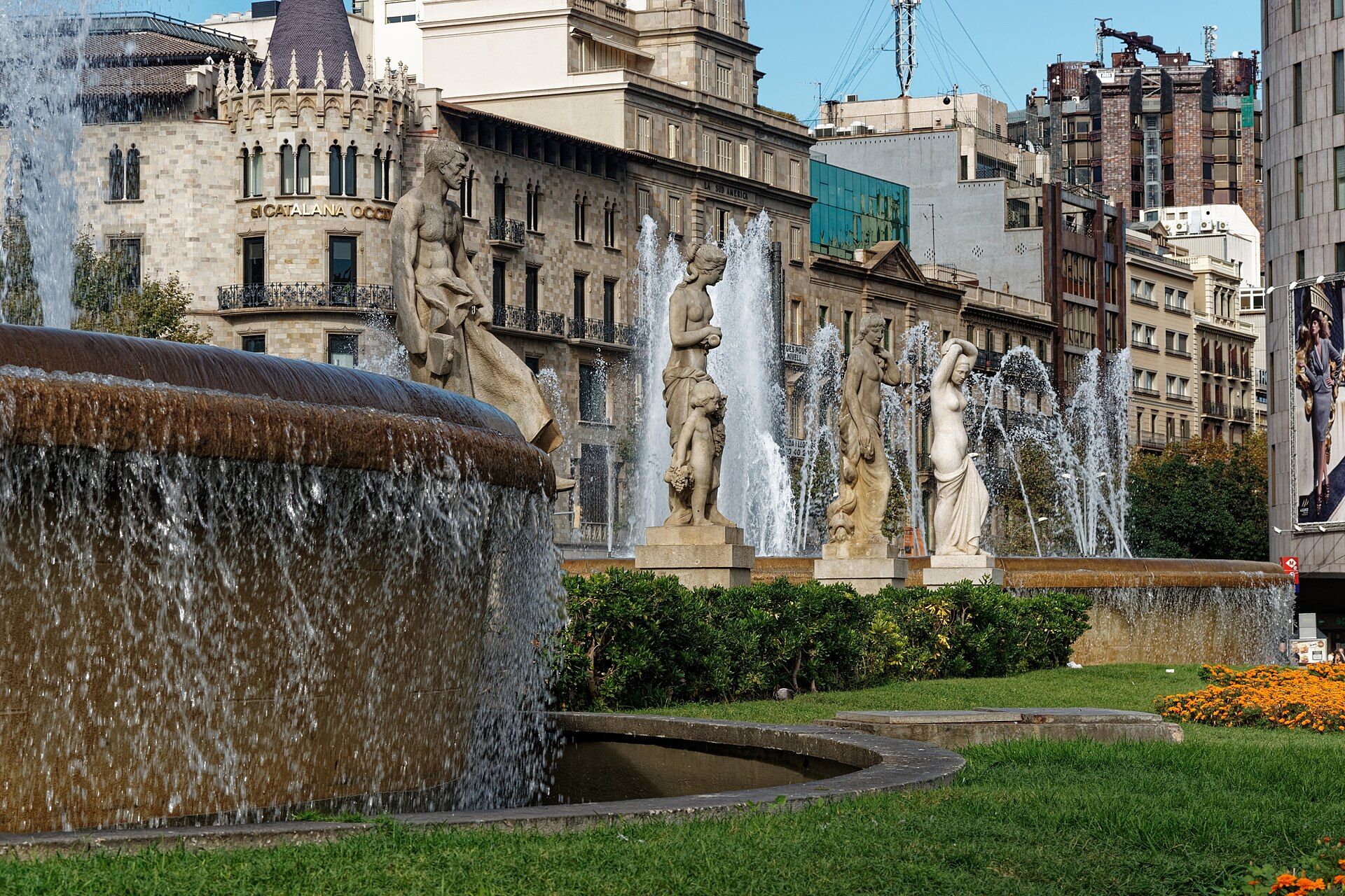 Fountain with stone statues on Plaça de Catalunya, with old office buildings behind on a sunny day.